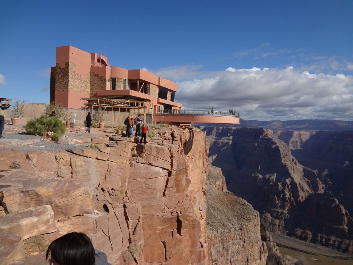 A pink-brown building stands at the edge of a red rock canyon, with a few visitors near the rim and a deep canyon stretching away under a blue sky with clouds.